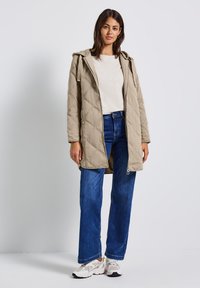 Gequilted, lichtbeige parka met een capuchon; beschikt over een ritsluiting en zijzakken. Gecombineerd met blauwe rechttoe rechtaan jeans en witte sneakers.