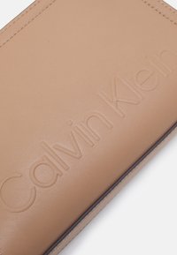 Bežs ādas maks ar iegravētu "Calvin Klein" logo, gludu tekstūru un izsmalcinātu šuvju līniju ap malām.