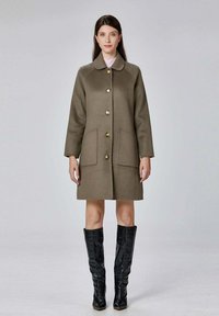 Manteau en laine vert olive avec un col, boutons dorés et deux poches avant. Porté sur un haut rose clair et assorti avec des bottes noires montantes.