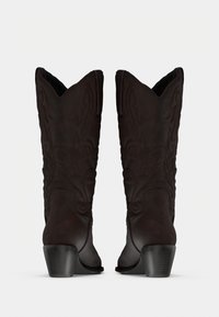 Ζευγάρι από σκούρα καφέ δερμάτινες καουμπόικες μπότες με κεντημένες λεπτομέρειες και στοίβες τακουνιών, απεικονιζόμενες από πίσω σε λευκό φόντο.