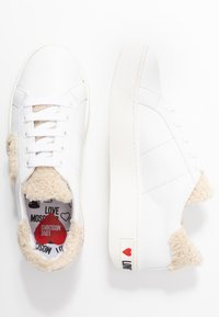 Vita lädersneakers med fluffiga beige detaljer, texturerad vit sula och ett mönstrat inre. Röd branding på hälen.