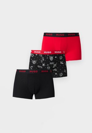 Ung mand står barfodet på tæppe iført sorte boxershorts med rød "HUGO" logo linning mod en ensfarvet lys baggrund.