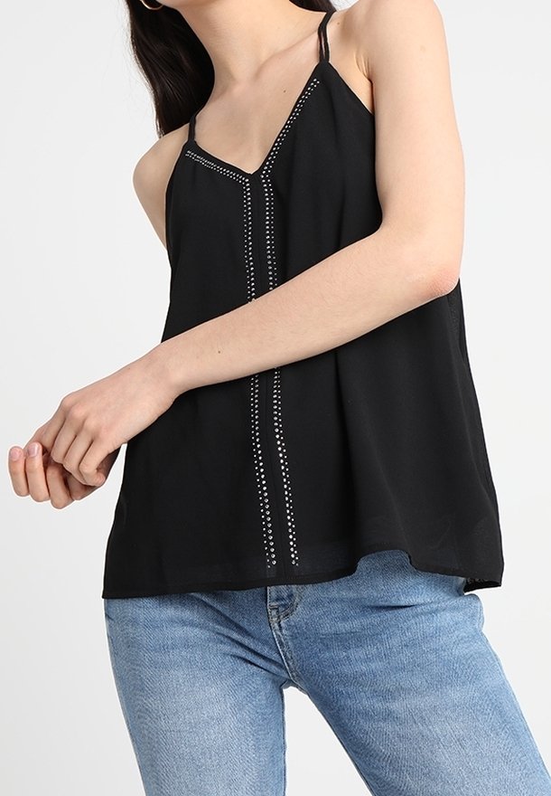 Femme portant un haut noir sans manches avec des clous verticaux décoratifs, associé à un jean bleu clair sur un fond uni.
