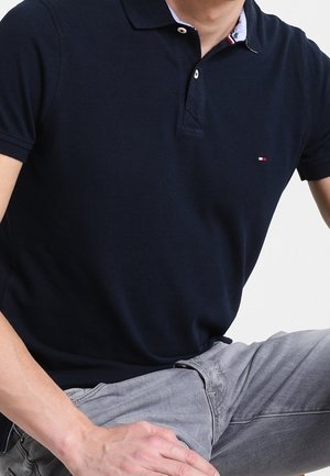 Mann trägt ein dunkelblaues Poloshirt mit kleinem rot-weiß-blauem Logo und hellgrauer Hose, sitzt mit den Händen auf den Oberschenkeln.