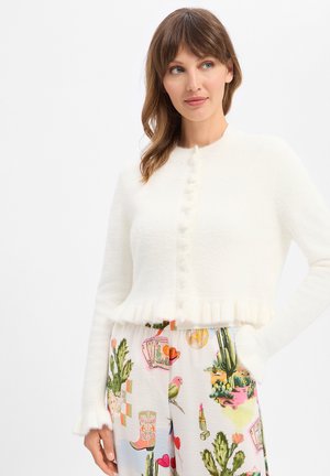 Frau mit braunen Haaren, die einen weißen, flauschigen, geknöpften Pullover und bunte Hosen mit Kaktus-, Vogel-, Lippenstift- und Cowboystiefelmuster trägt.