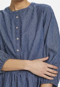 Žena ve modré strukturované blůze s knoflíky a 3/4 rukávy, držící svou levou ruku pravou rukou na úrovni pasu.