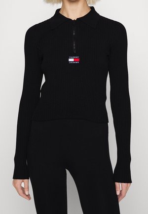 Kvinde iført en sort ribbet langærmet top med lynlåshals og Tommy Jeans-logo på brystet, kombineret med sorte bukser.