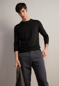 Maglietta nera a maniche lunghe con una texture liscia e scollo rotondo, abbinata a pantaloni grigi su misura. Il modello tiene una giacca scura in una mano.