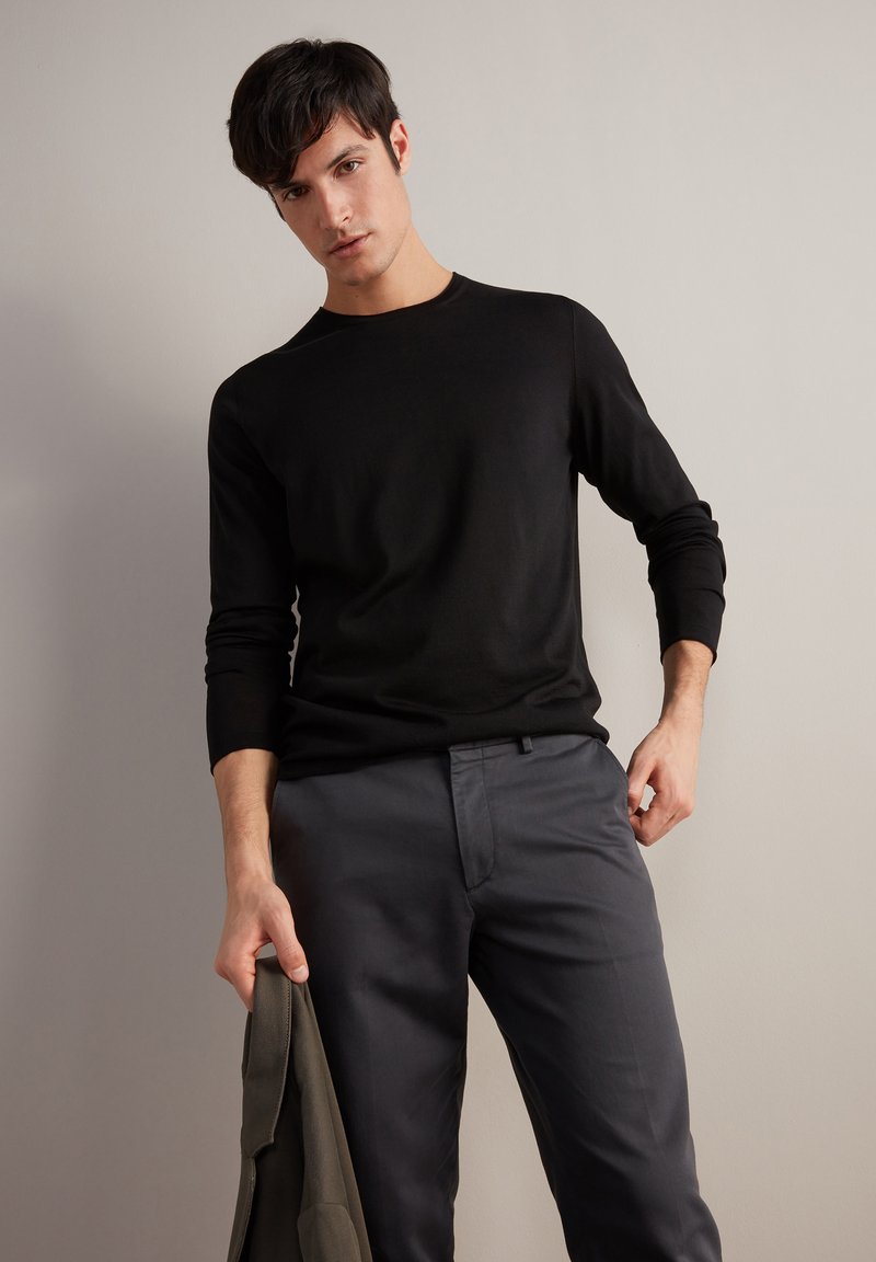 Maglietta nera a maniche lunghe con una texture liscia e scollo rotondo, abbinata a pantaloni grigi su misura. Il modello tiene una giacca scura in una mano.