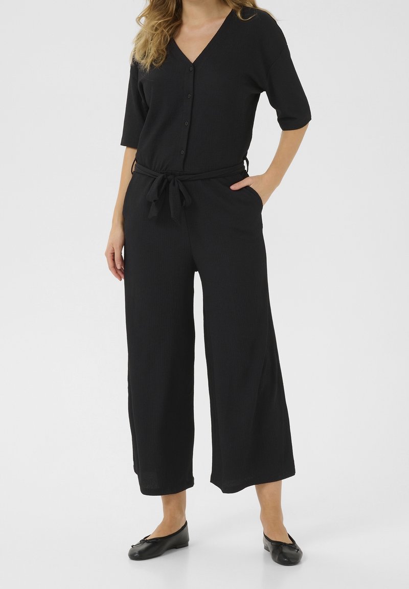 Femme portant un haut noir boutonné avec manches trois-quarts et un pantalon noir à jambes larges avec une ceinture nouée à la taille ainsi que des chaussures plates noires.