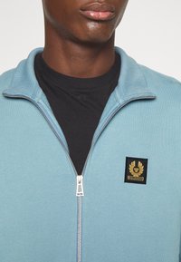 Chaqueta de cremallera azul claro con cuello acanalado y detalles en la cremallera, con un parche negro con un emblema dorado en el lado izquierdo del pecho, sobre una camiseta negra.