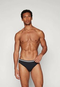 Uomo muscoloso con capelli corti e ricci, che indossa slip neri con strisce bianche in vita, in piedi contro uno sfondo grigio uniforme.