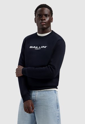 Man met over elkaar geslagen armen, gekleed in een zwart sweatshirt met de tekst "BALLIN'" en lichtblauwe spijkerbroek, staande tegen een effen grijze achtergrond.