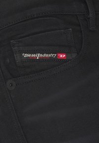 Black denim jeans with a small pocket featuring a red and silver "Diesel Industry" label and a black button. Smooth texture, slim fit design.