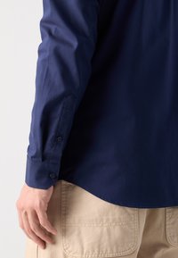 Cămașă bleu marin cu manșete cu nasturi, având o textură netedă și un tiv ușor curbat, asociată cu pantaloni bej deschis, dotati cu buzunare.