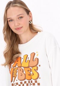 Felpa bianca con grafiche "FALL VIBES" in tonalità arancioni e marroni; presenta un motivo a scacchi e un collo rotondo. I capelli sono mossi.