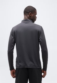 Άνδρας που κοιτάζει προς τα πίσω, φορώντας εφαρμοστή μακριά μπλούζα σε σκούρο γκρι χρώμα και μαύρο παντελόνι μπροστά σε απλό ανοιχτό φόντο.