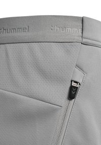 Close-up van grijs sportkledingstof met een reliëf "hummel" logo op de tailleband en een verticale rits met een zwart trekkoord aan de zijkant.