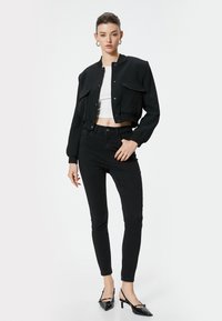 Chaqueta bomber negra corta con botones a presión y dos bolsillos delanteros, combinada con jeans negros ajustados y zapatos de tacón negros puntiagudos.