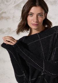 Schwarzer, strukturierter Pullover mit lockerer Passform, der ein Gittermuster aus weißen Nähten und weiten Ärmeln aufweist. Weicher Stoff mit einem gemütlichen Aussehen.