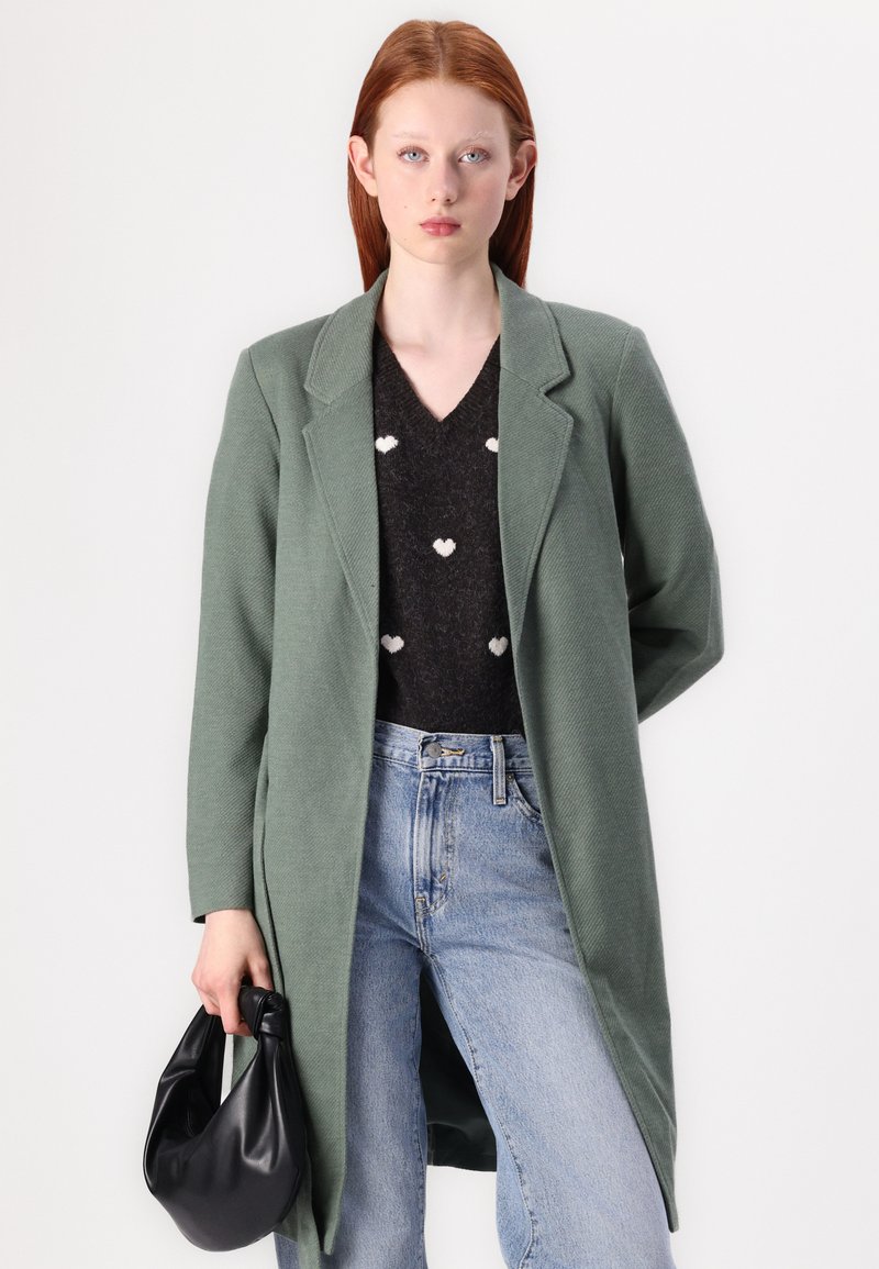 Grüner langer Mantel mit maßgeschneidem Design, aus strukturiertem Stoff, getragen über einem dunklen Pullover mit Herzmustern, kombiniert mit hellblauen Jeans und einer schwarzen Tasche.
