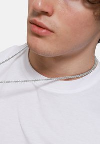 Giovane che indossa una collana d'argento sopra una maglietta bianca a girocollo, con un primo piano su labbra e linea della mascella.