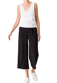 Witte mouwloze V-hals top gecombineerd met zwarte wijde broek met een trekkoord in de taille, en afgewerkt met witte sneakers.