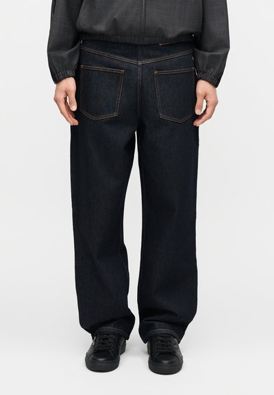 Άνδρας που φοράει φαρδιά σκούρα μπλε τζιν παντελόνια και μαύρα αθλητικά παπούτσια, εμφανιζόμενος από πίσω με τα χέρια χαλαρά στο πλάι.