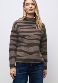 Brązowy, puszysty sweter z ciemnymi paskami, wysoki kołnierz i długie rękawy, zestawiony z niebieskimi dżinsami. Miękka tekstura i luźny krój.