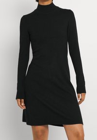 Svart långärmad klänning med hög halsringning, tillverkad av mjukt tyg. Den har en avslappnad passform och sidoöppningar nära nederkanten. Slät textur.