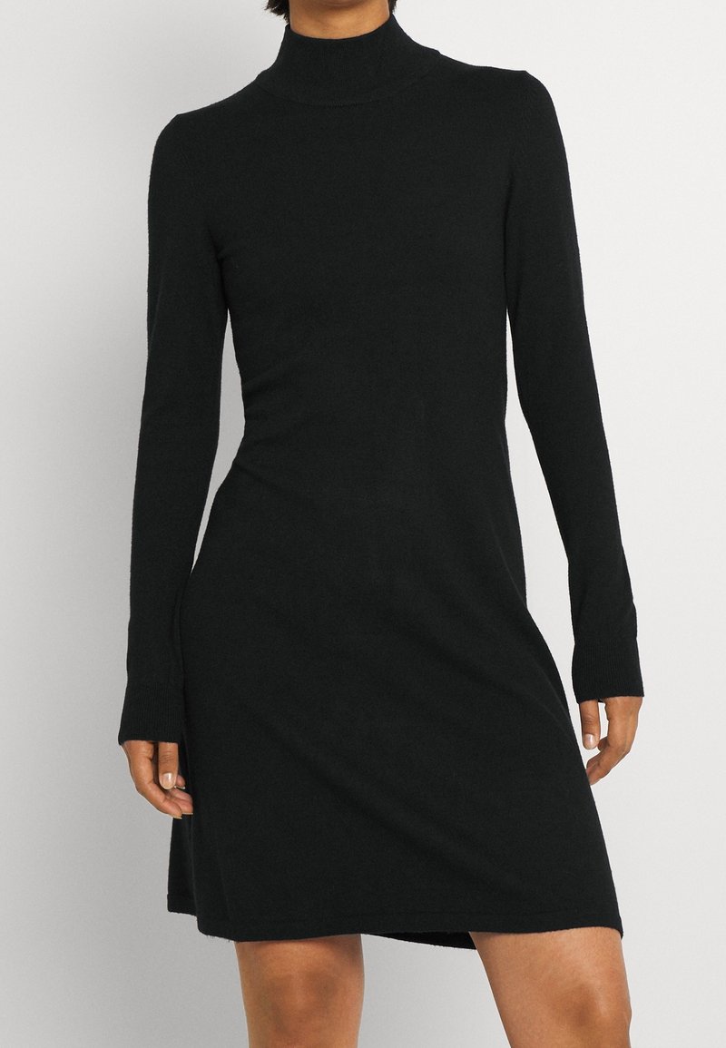 Svart långärmad klänning med hög halsringning, tillverkad av mjukt tyg. Den har en avslappnad passform och sidoöppningar nära nederkanten. Slät textur.