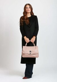 Borsa a tracolla in pelle rosa con due fermagli argentati, texture morbida e forma strutturata. Indossata con un lungo cappotto nero e jeans scuri.