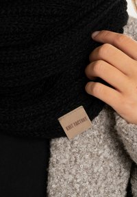Zwarte gebreide sjaal met een gestructureerd patroon en een beige rechthoekig label met "Knit Factory" in bruine letters, gedragen tegen een lichtgrijze jas.