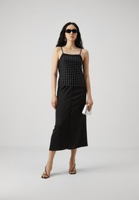 Haut sans manches noir avec des clous métalliques, associé à une jupe midi noire. Les accessoires comprennent une pochette argentée et des sandales noires à lanières.