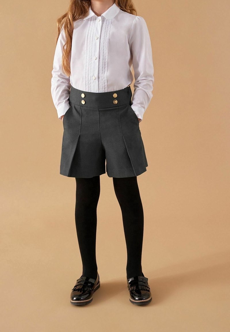 Enfant portant une chemise blanche à manches longues boutonnée, un short taille haute foncé avec des boutons dorés, des collants noirs et des chaussures noires brillantes, debout les mains dans les poches.