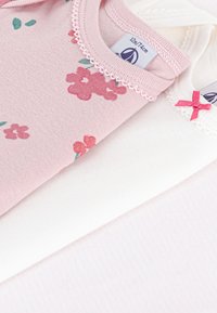 To bomullsbodyer – en rosa med blomstertrykk og blonder, den andre hvit med en liten rosa sløyfe og blonde detaljer. Myke teksturer.
