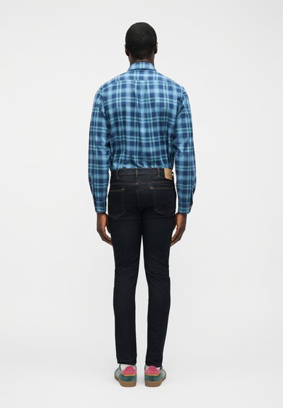 Άνδρας που στέκεται με την πλάτη προς τα εμάς, φοράει μπλε καρό μακρυμάνικο πουκάμισο, σκούρο τζιν και πολύχρωμα αθλητικά παπούτσια σε απλό φόντο.