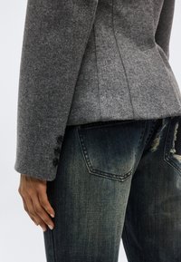 Άτομο που φοράει ένα εφαρμοστό γκρι μάλλινο σακάκι με τρία μαύρα κουμπιά στη μανσέτα και σκούρο ξεβαμμένο τζιν.