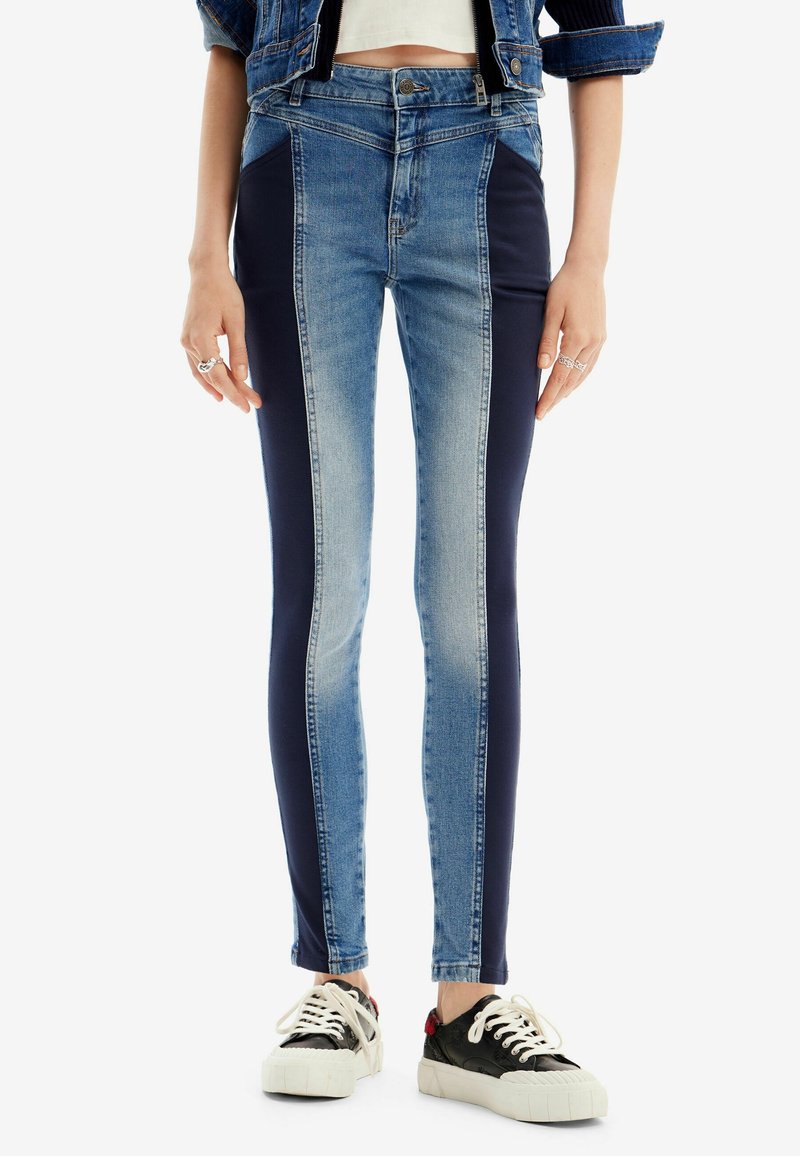 Personne portant un jean skinny taille haute avec des panneaux latéraux foncés, une veste en jean courte, un top blanc court et des baskets noir et blanc.