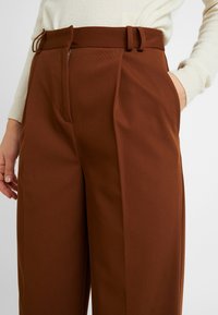 Bruna högmidjade byxor med texturerad yta, med veck, bälte och sidofickor. Materialet ser vävt och strukturerat ut.