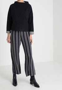 Svart textured tröja med vid halsringning och långa ärmar, tillsammans med svart och vitrandiga vida byxor och svarta ankelskor.