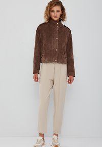 Bruin suede cropped jasje met een hoge kraag, voorzakken en gouden knopen, gecombineerd met beige broeken en beige sneakers.