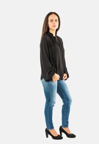 Femme se tenant de profil, portant un blouse noire ample, un jean bleu ajusté et des chaussures à talons noirs sur un fond uni.