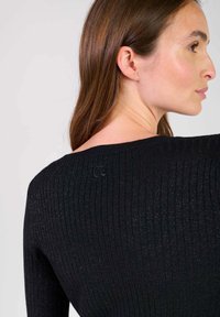 Pull en maille noire à côtes avec un large col et un éclat subtil. Arrière orné d'un petit logo. Le matériau semble doux et texturé.