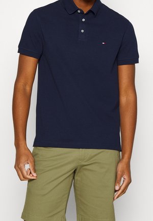 Homme portant un polo bleu marine à manches courtes avec un logo sur la poitrine et un short vert olive longueur genou, debout devant un fond uni.