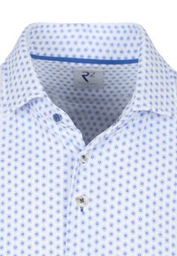 Witte overhem met een cirkelvormig blauw patroon, voorzien van een blauwe voering bij de kraag en twee knopen zichtbaar in het halsgebied.