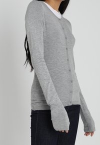 Žena v upnutém šedém cardigan u s knoflíky, oblečená přes bílou košili a tmavé džíny, stojící na pozadí s jednobarevným světlým pozadím.
