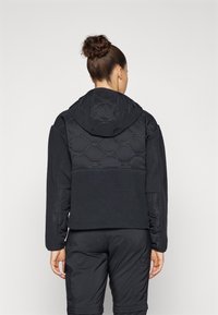 Chaqueta negra con capucha acolchada, mangas de forro polar texturizado y un diseño ajustado. Presenta patrones circulares en el panel trasero.