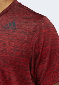 Camiseta atlética roja con un patrón texturizado, escote de cuello redondo y mangas cortas. Presenta el logo negro de adidas en la parte superior izquierda.