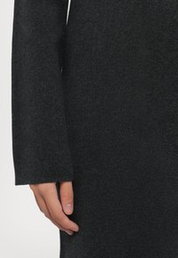 Sötét szürke pulóver bordázott textúrával és hosszú ujjal, egyenes vonalú szabással. A kéz, amely a szöveten nyugszik, a puhaságot tükrözi.